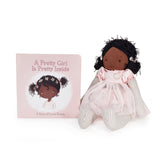 A soft fabric doll with dark brown skin and curly black hair stands beside a pink children's book titled "A Pretty Girl Is Pretty Inside" on a clean white background.