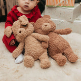 Child in a red outfit holding two brown plush animals on a light-colored surface.