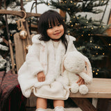 Child in a white fluffy coat holding a plush toy in front of a decorated Christmas tree.
