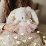 A white plush bunny with long floppy ears and a pink tulle tutu sits on a child's lap. The child is wearing a pink floral dress, and small white daisies are scattered in the foreground.