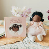 A children's book titled "A Pretty Girl Is Pretty Inside" stands next to a soft fabric doll with dark curly hair, a pale pink dress, and striped legs on a floral tablecloth with flowers in the background.