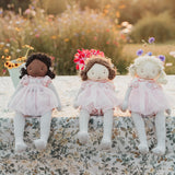 Three handcraftedcloth dolls with striped socks and pink dresses sit side by side on a floral tablecloth in an outdoor garden setting with warm golden-hour sunlight.