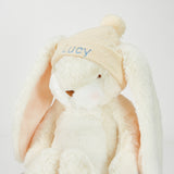 Plush bunny toy with a beige knit hat labeled 'Lucy' on a light gray background