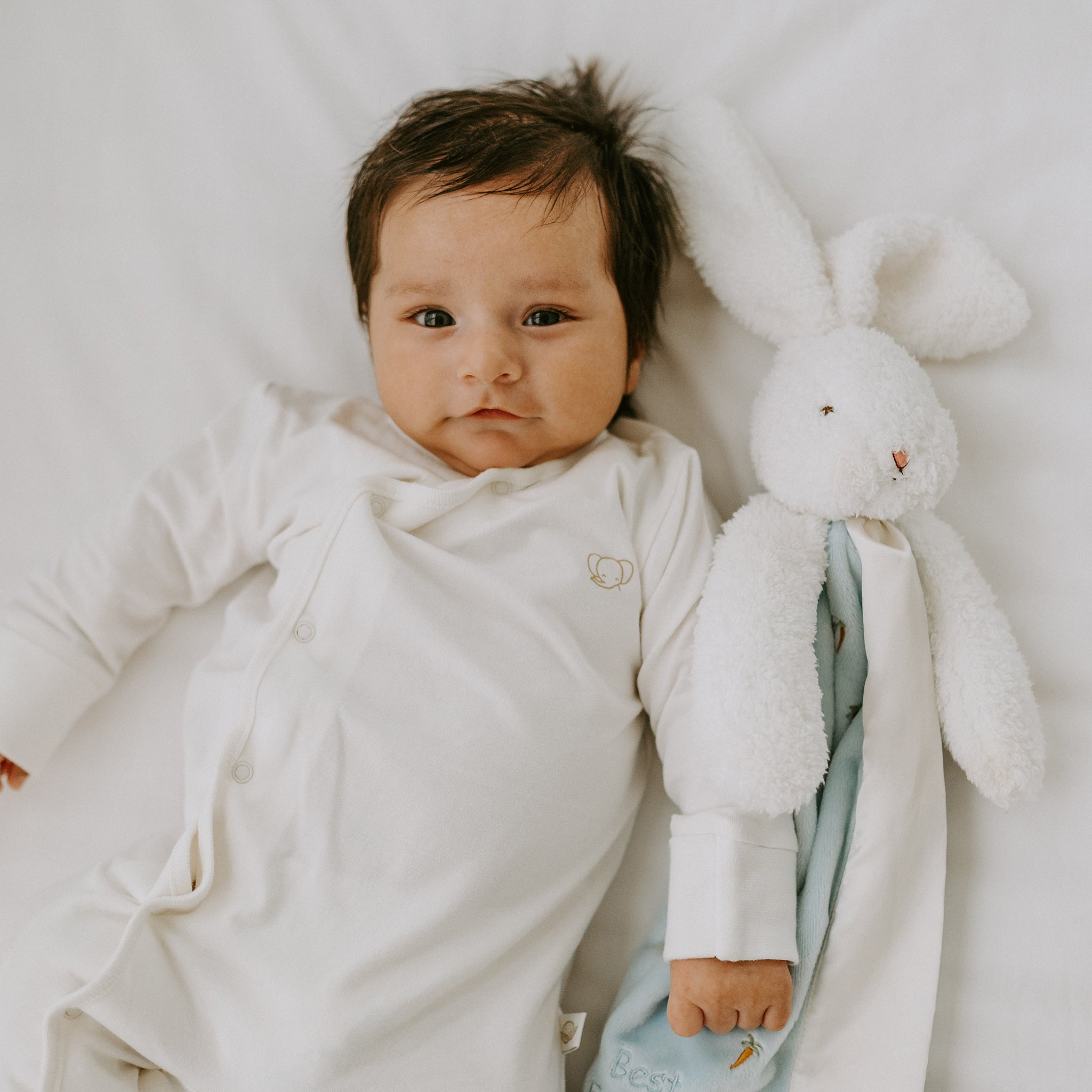 Baby in a white onesie lying next to a plush bunny buddy blanket toy on a soft surface