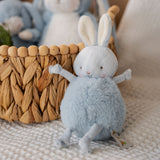 A small plush toy resembling a round blue bunny with a velour face, dangly arms and legs, and embroidered features, sitting on a white blanket with a woven basket next to it.