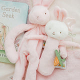 Pink plush bunny and pink bunny rattle with carrot tag on a pink blanket next to a children's book