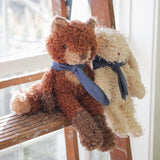 A fox and lamb stuffed animal, one brown and one beige, sitting on a wooden surface with a blurred background.