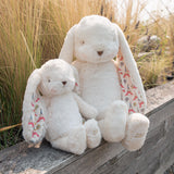 Two white plush bunny toys with long floppy ears sit on a weathered wooden bench outdoors, with tall dry grass in the background. Each bunny has patterned inner ears featuring colorful Eiffel Tower and Paris-themed motifs.