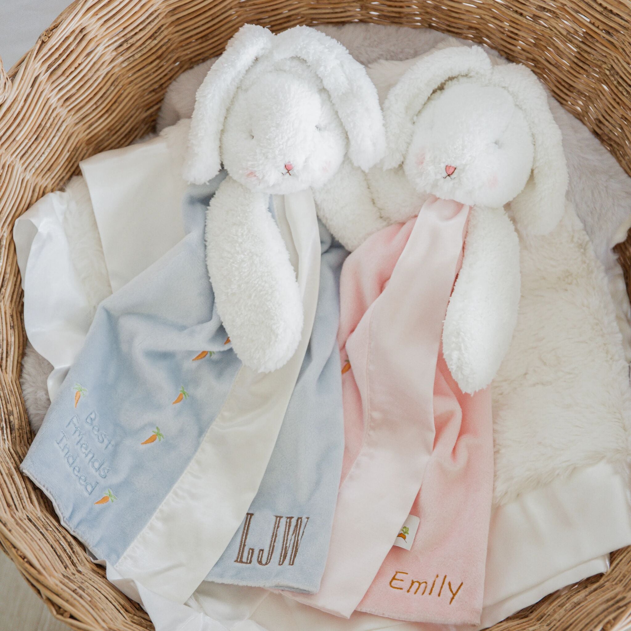 Lifestyle shot of Two white plush bunny buddy blanket toys with custom embroidered names styled in a woven basket.