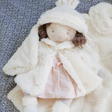 Brown haired doll in a pink dress and a cream fuzzy coat and hat, laying on a fuzzy background.