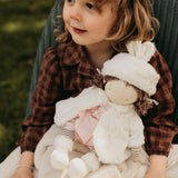 Child holding a doll in a cream colored fuzzy jacket and hat with bunny ears.