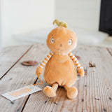 A plush toy doll with a round orange body, striped yellow and white arms, and a small green leaf on its head, sitting on a rustic wooden table with a small tag attached.