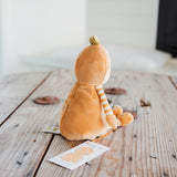A small plush toy shaped like a round orange character with a striped arm and a small green top, shown from the back on a rustic wooden table with a tag in front.