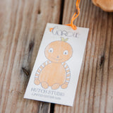 A small white paper hang tag with an orange string, featuring a cartoon pumpkin character and the text 'Gordie' and 'Hutch Studio Limited Edition 2026, photographed on a rustic wooden surface.