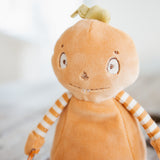 A close-up of a plush toy with a round orange-brown body, striped orange-and-white arms, and a small green leaf on top, photographed against a soft blurred light background.