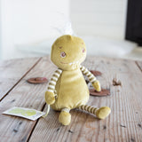A small green plush monster toy with striped arms and legs, sitting on a rustic wooden table. The toy has embroidered eyes and mouth, a small white tuft on its head, and a small paper tag attached near its hand.