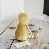 A green plush stuffed animal toy with a white tuft on its head and striped legs, shown from the back on a rustic wooden table.