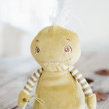 A handmade green plush monster toy with striped tuft, embroidered eyes, and a white feather on its head, photographed in a soft, bright setting.