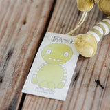 A plush toy with a green monster face and striped olive-green and cream body, resting on a rustic wooden surface next to a paper tag labeled 'FRANKIE' and with'Hutch Studio' branding.