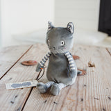A gray plush cat toy with striped arms and legs, sitting on a rustic wooden table. The toy has embroidered facial features and a small paper tag attached to its wrist.
