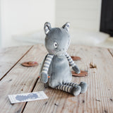 A gray plush cat toy with striped arms and legs sits on a rustic wooden table. A small printed card is placed near the toy, and the background shows a softly blurred indoor setting.