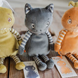 Three plush stuffed toys arranged on a wooden surface: a gray cat in the center with striped arms and legs, a green monster on the left, and an orange pumpkin-like character on the right. Each toy has a paper tag attached.