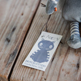 A close-up of a plush gray stuffed animal with a striped leg lying on a rustic wooden surface next to a paper tag printed with the product name 'Scaredy' and a cat illustration.