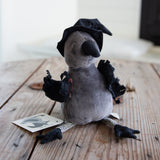 A plush toy bird dressed as a witch, sitting on a rustic wooden surface. The toy features a gray body, a black pointed hat, black tattered wings, and striped black-and-white legs.