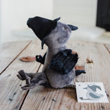 A small plush crow doll with a gray body, black hat and wings, sitting on a rustic wooden surface. A small printed card with a witch illustration is placed beside it.