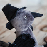 A close-up of a small plush toy with a gray body, black pointed hat, and black wings, photographed on a softly blurred wooden surface.
