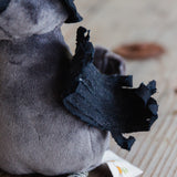 Close-up of a gray plush toy with a black sewn fabric wing attached to its side, resting on a light wood surface.