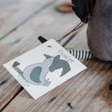 A close-up of a handmade plush toy with a white paper tag that reads 'CROWLEY' and shows a gray and black animal illustration. The tag is attached to a striped tail, and the plush is next to a gray fabric body on a rustic wooden surface.
