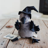A small plush toy bird dressed as a witch, sitting on a rustic wooden surface. The toy has a gray body, a black felt beak, a black pointed witch hat, and black ruffled wings and feet.