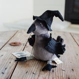 A small plush toy bird dressed as a witch, sitting on a rustic wooden surface. The toy has a gray body, a black pointed hat, black wings, and striped black-and-white legs.