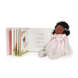 A children's book with a plush doll beside it on a clean white background. The book is open to a poem page with illustrations, and the doll has dark curly hair, a pink dress, and striped pink-and-white legs.
