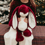 Person holding a plush bunny with red ears and scarf in front of Christmas trees.