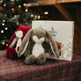 Plush bunnies by a gift box on a plaid surface with a blurred Christmas tree in the background.