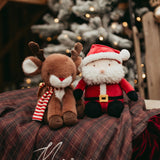 Two Christmas-themed plush toys, a reindeer and Santa Claus, on a plaid surface with a blurred festive background.