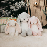 Three plush long-eared bunny toys in white, light gray, and light pink are arranged on a white faux-fur surface with a Christmas tree, wooden fence, and gift box in the background.