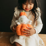 A young child in a cream knit dress holds a plush toy shaped like a white bunny inside an orange pumpkin, posed against a dark background.