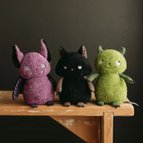 Three fuzzy plush toy halloween creatures sitting side by side on a wooden table against a dark background. The left toy is purple with black ears and purple striped wings. black kitty cat in the middle, and green monster on the right. 