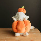 A plush toy featuring a white sheep nestled inside a large orange pumpkin, with a smaller orange pumpkin on its head, sitting on a wooden surface against a dark background.