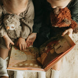 Two children reading a book together, surrounded by cozy clothing and a plush owl toy.
