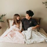 Two children sitting on a bed with pink and gray blankets, surrounded by plants.