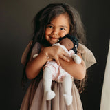 A young girl with long dark hair holds a doll dressed in a light pink outfit and white socks against a dark studio background.