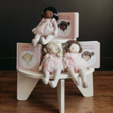 Three cloth dolls in pink dresses posed on a small white stool with matching book covers behind them. The books and dolls are arranged on a wooden floor against a dark wall.