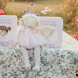 A soft fabric doll in a pink dress and striped tights sits on a floral tablecloth outdoors, with two copies of the children's book 'A Pretty Girl & is Pretty Inside' placed behind it. The background is a blurred warm-toned garden setting.