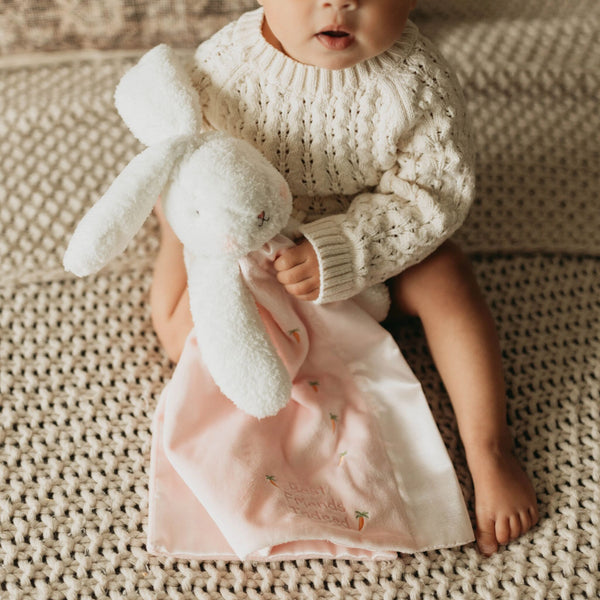 Baby holding a plush bunny toy and pink blanket on a textured surface