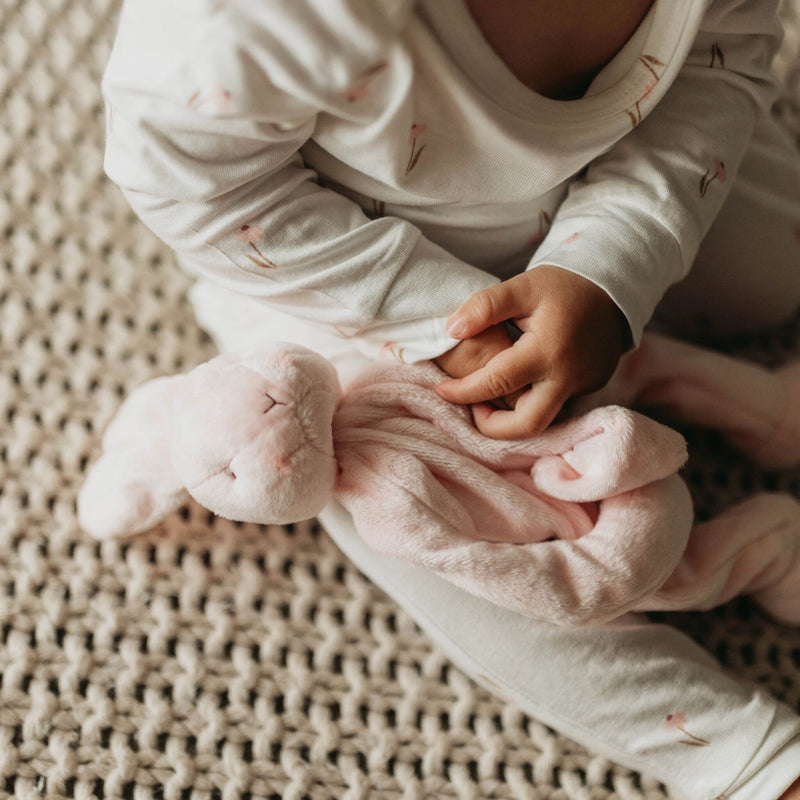 small Child in floral pajamas holding a pink plush bunny pacifier holder toy on a textured surface