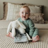 Baby in a green outfit holding a plush bunny on a textured surface
